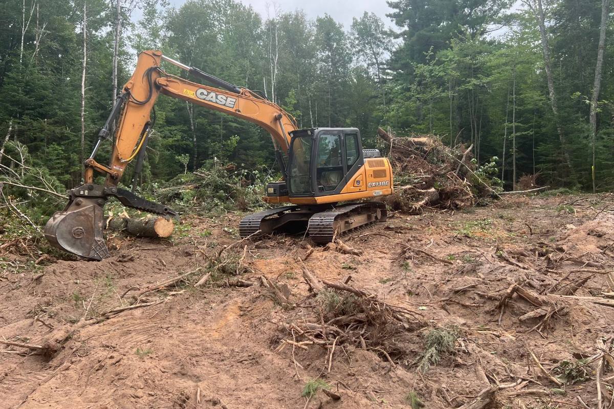 Excavator with a thumb clearing trees and brush from a lot, with the debris piled up behind